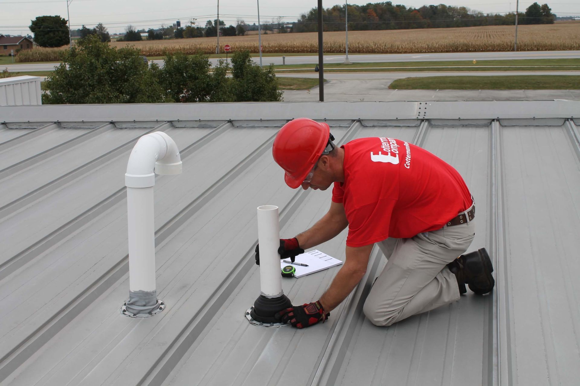 Man in red shirt and hard hat installs a vent pipe on a metal roof.