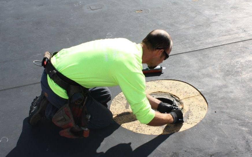 Person on roof repairs a circular opening, wearing safety gear and using a sealant gun.