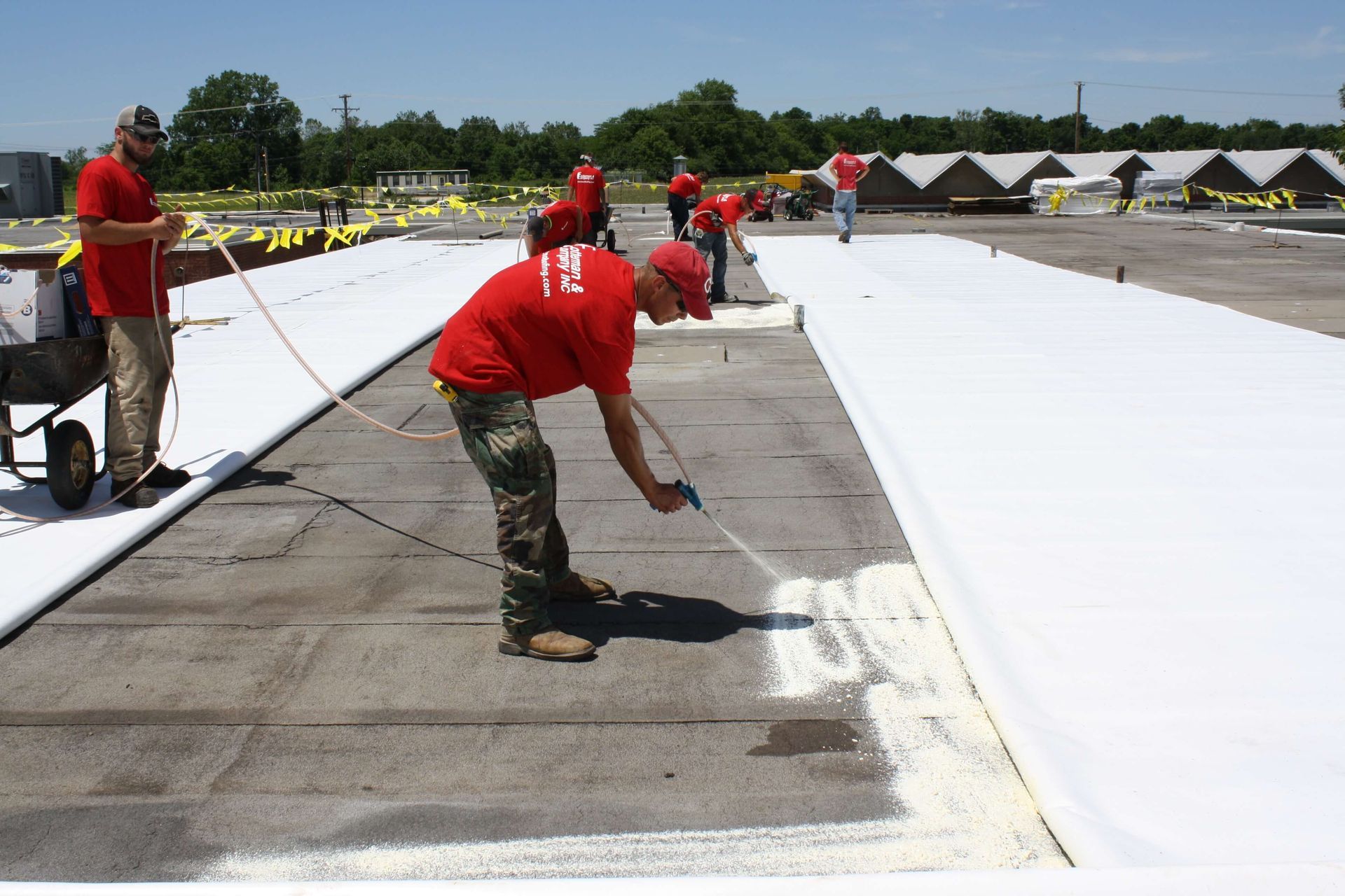 Workers in red shirts spray white sealant on a flat roof.