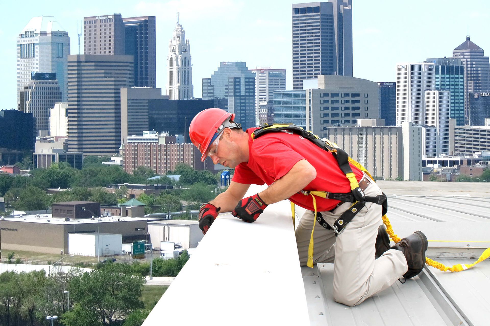 Construction worker on rooftop, wearing safety harness, inspecting edge with city skyline background.