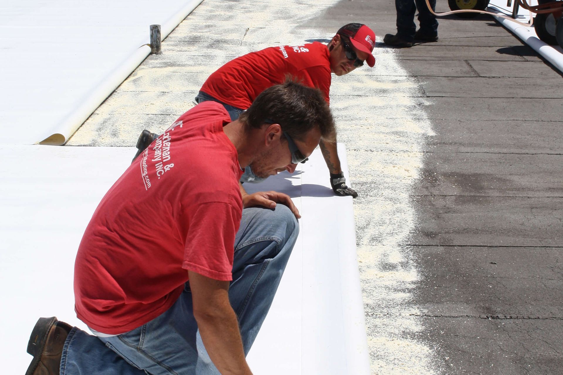 Two workers in red shirts installing white roofing material on a flat roof.