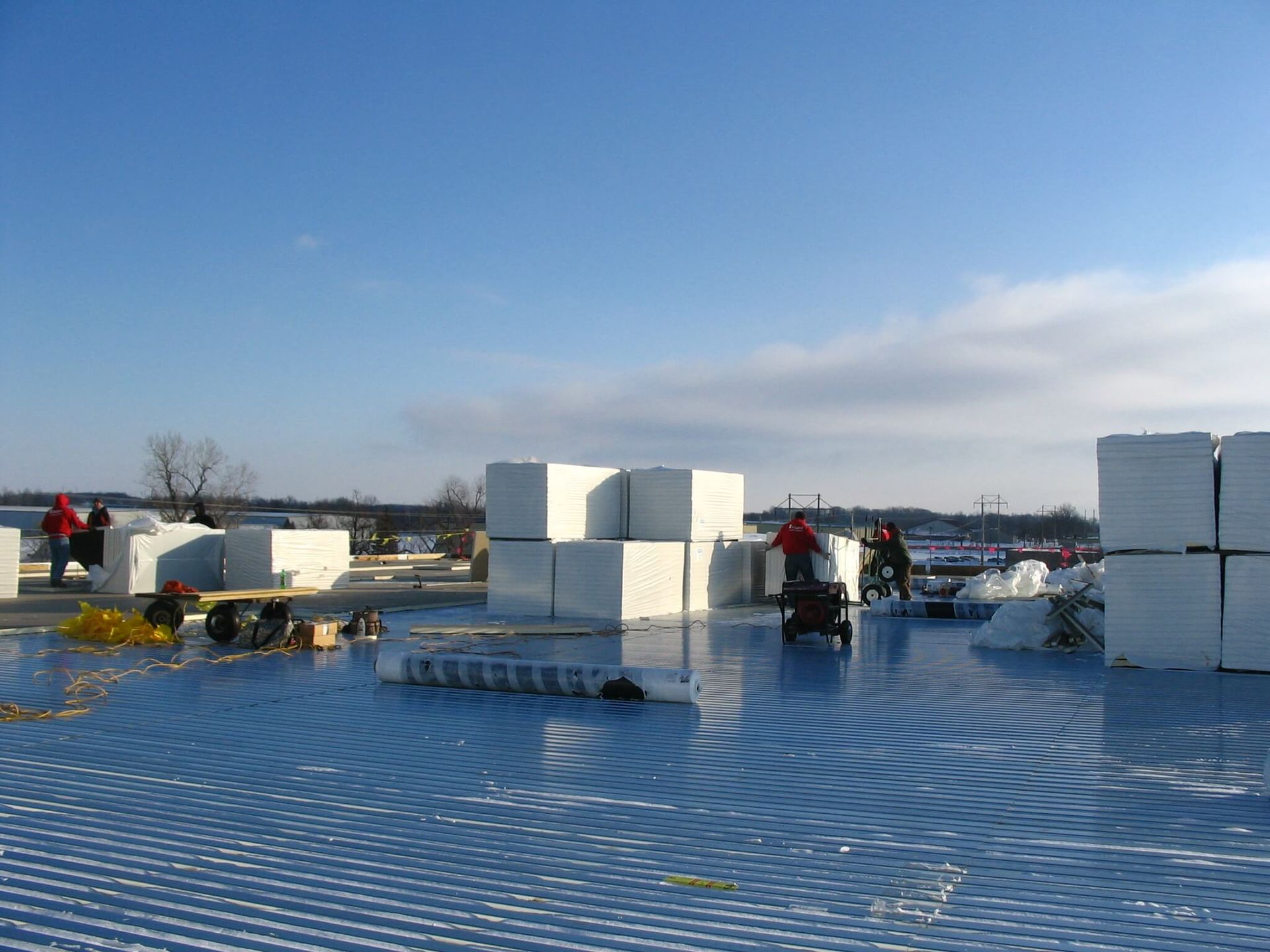 Workers on a roof installing insulation; blue membrane, white foam blocks, sunny day.
