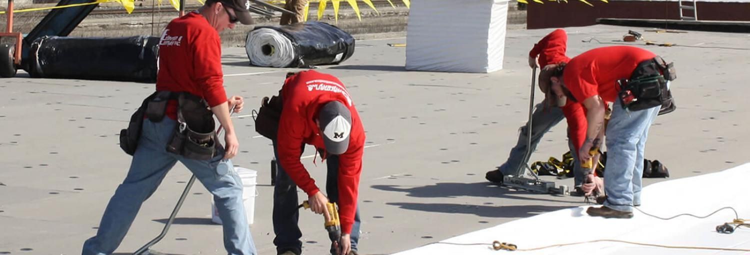 Roofing workers in red shirts and jeans on a rooftop. They're working on a white surface, with tools and materials nearby.