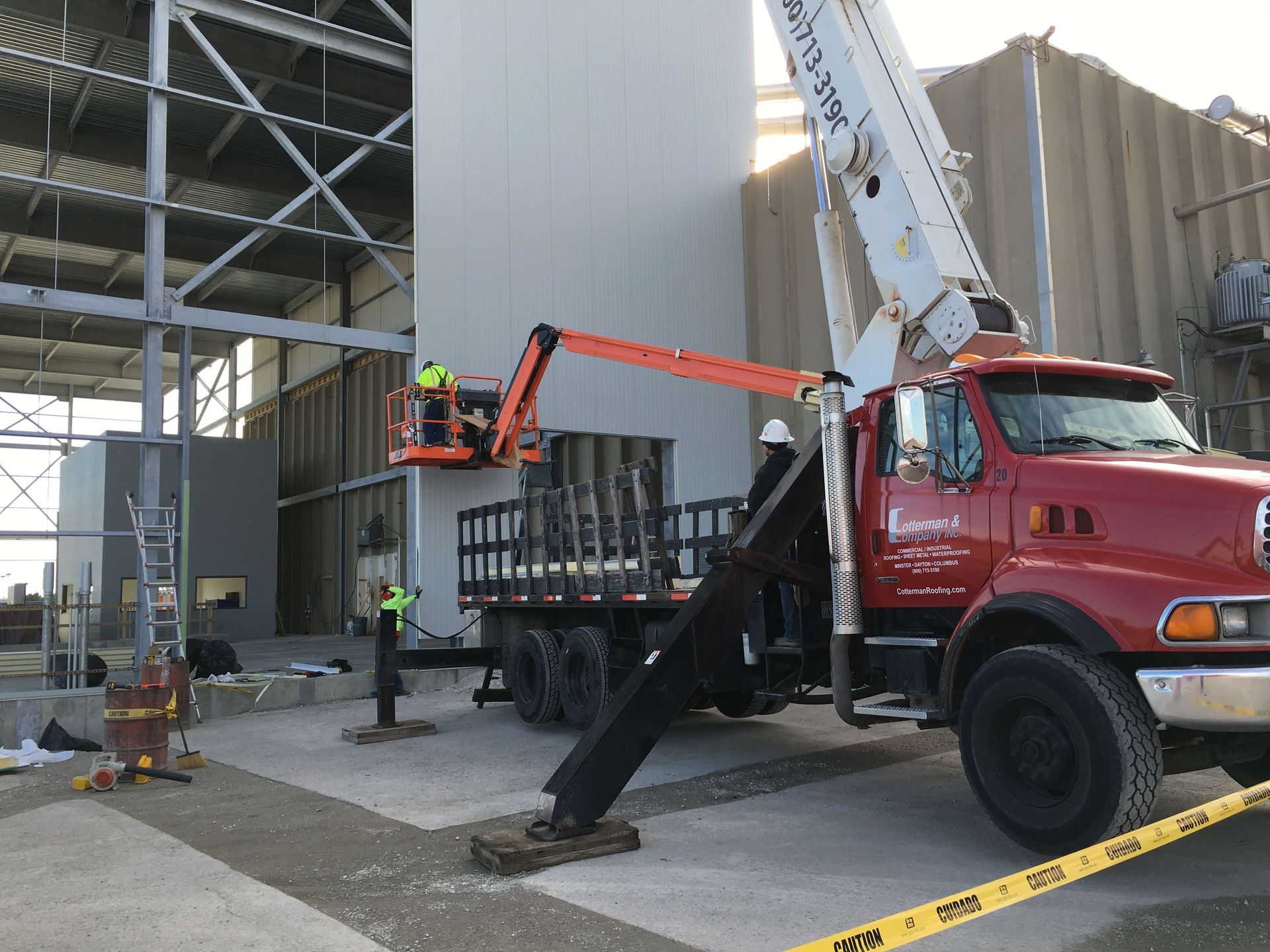 Workers on a boom lift loading cylindrical objects from a flatbed truck near a building.