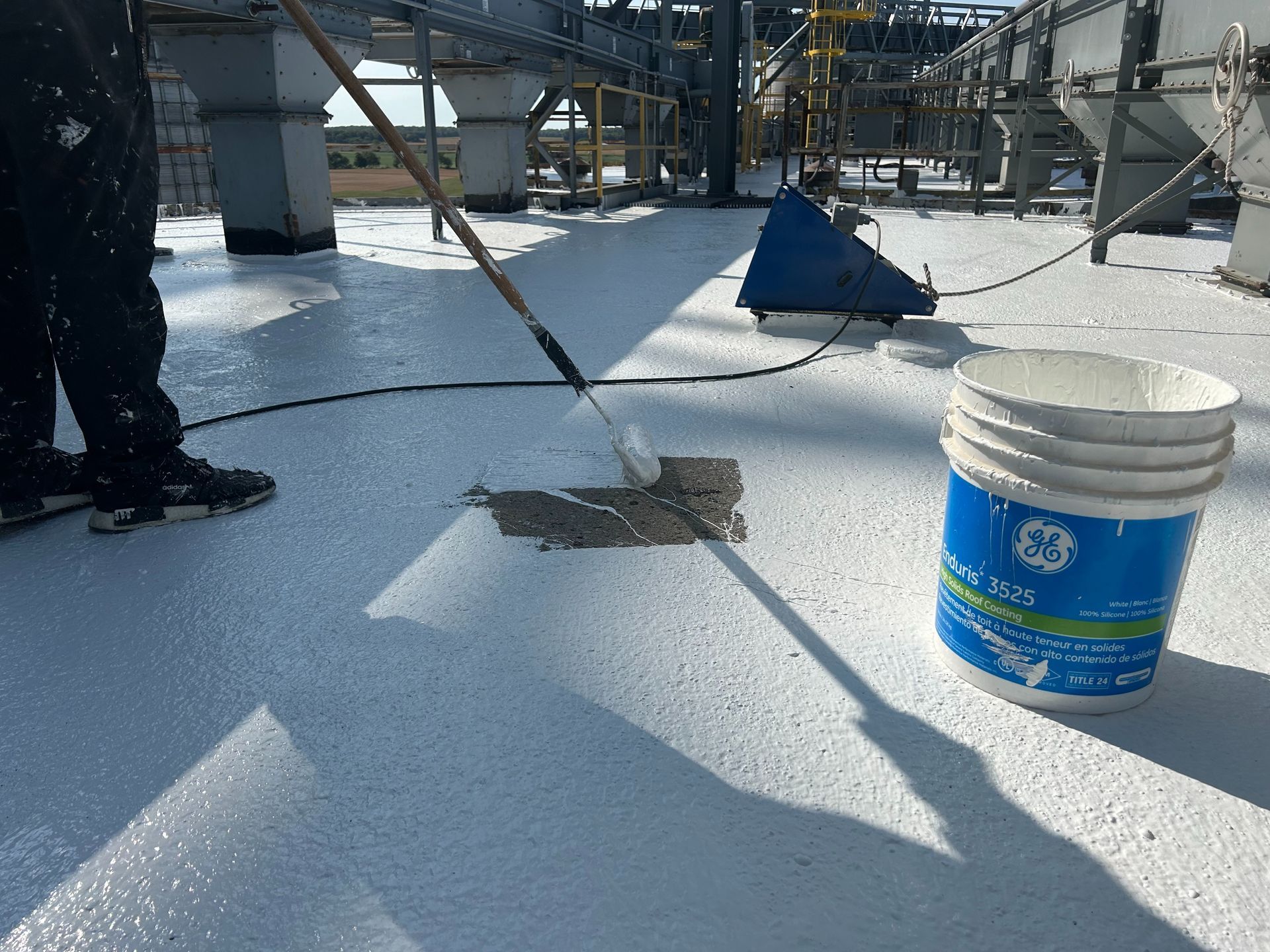 Person applying a white coating to a rooftop, with a bucket of the material nearby. Industrial setting.