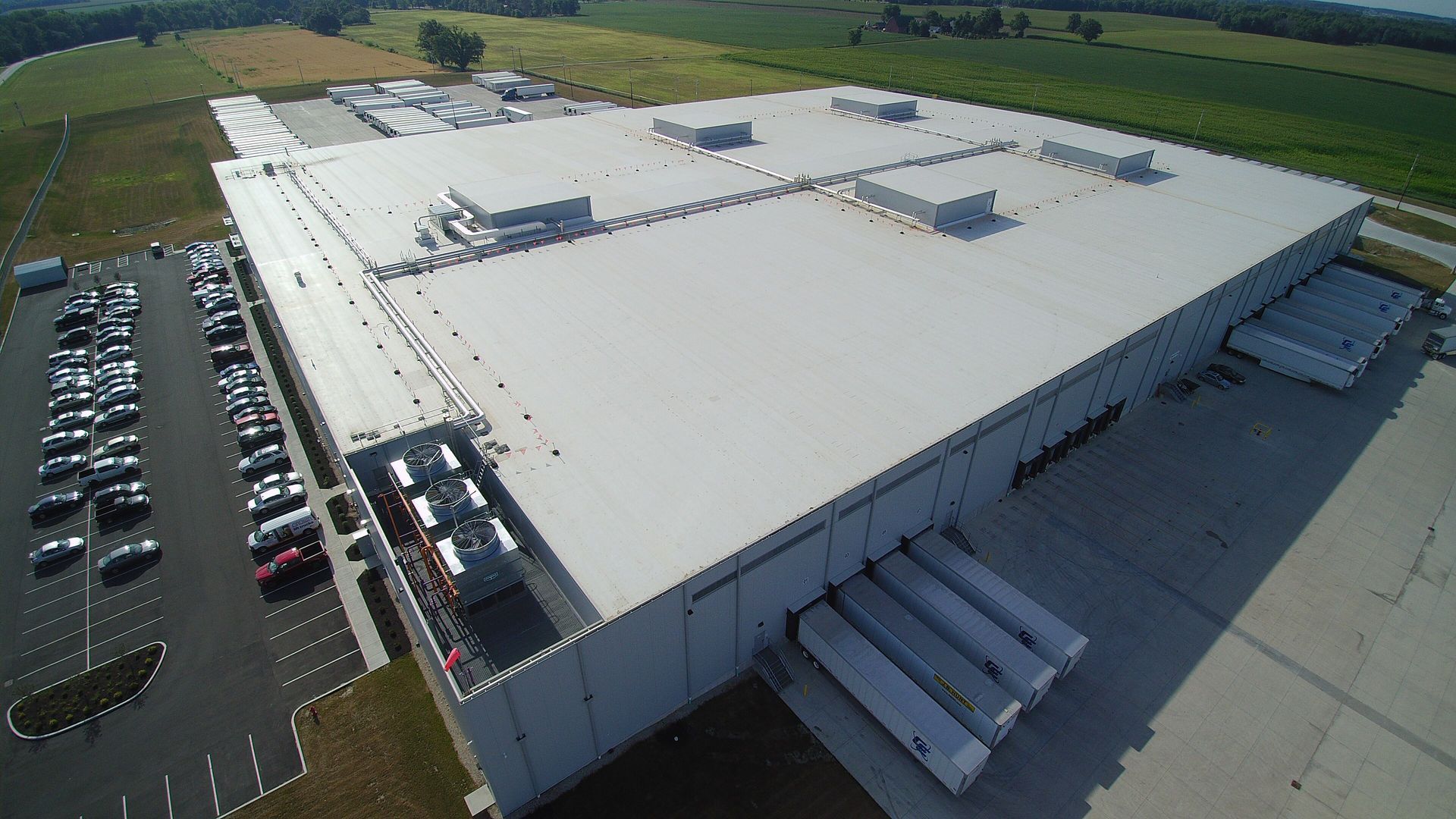 Aerial view of a large industrial building with a flat roof, loading docks, and a parking lot filled with vehicles.