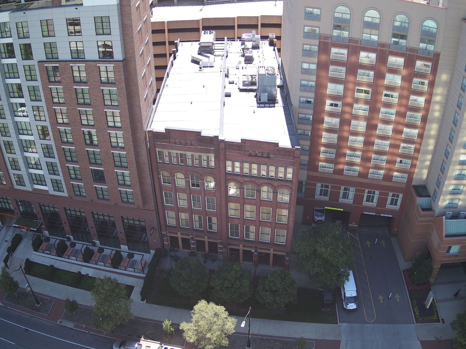 Buildings in a city: brick, modern, and concrete structures, with a white vehicle and green trees below.