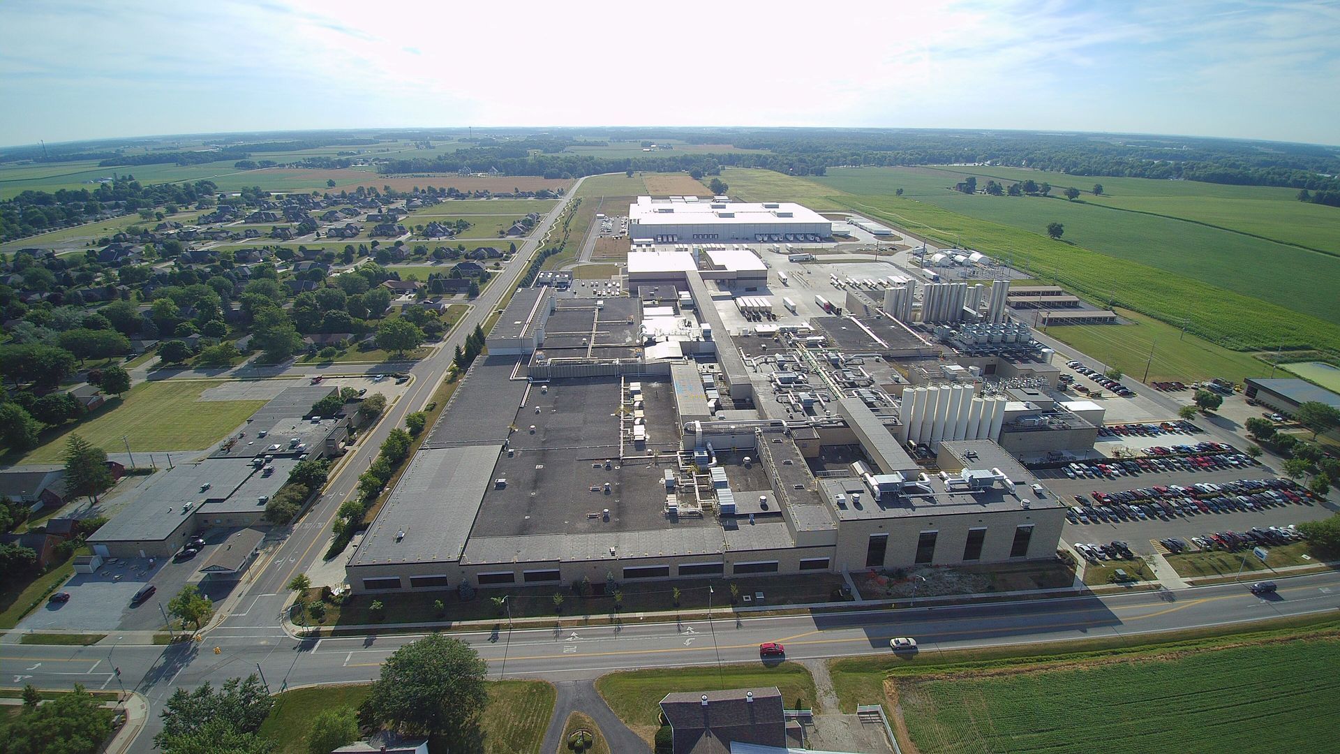 Aerial view of a large industrial complex with buildings, parking lots, and surrounding farmland under a blue sky.