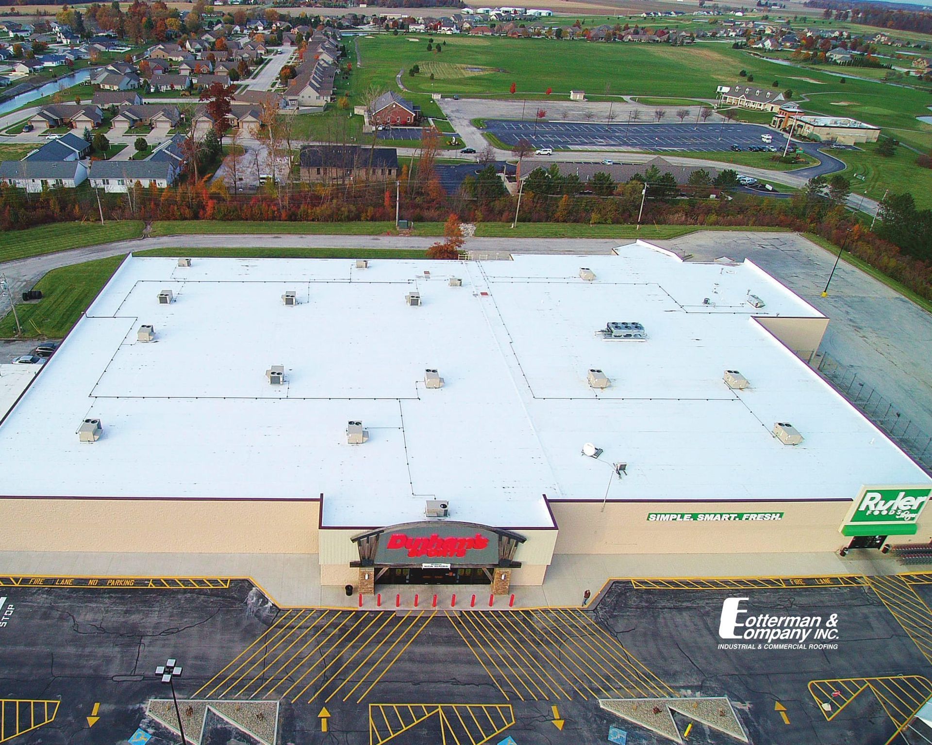 Aerial view of a large building with a white roof, possibly a store, with surrounding parking and residential areas.