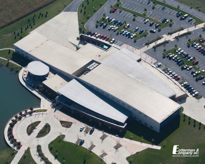Aerial view of modern building with long, angular roof, surrounded by parking and pond.