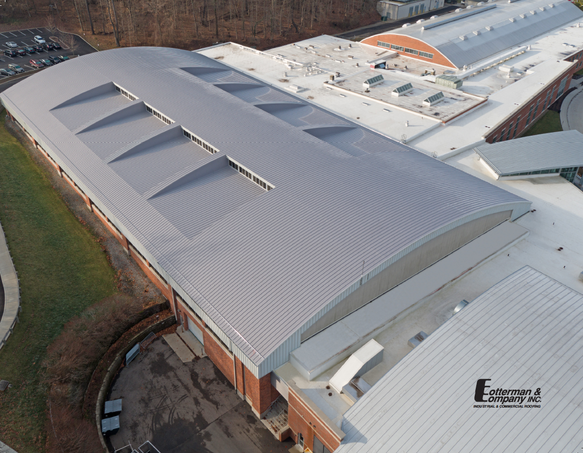 Aerial view of a long, curved roof with several skylights, made of light-colored metal.