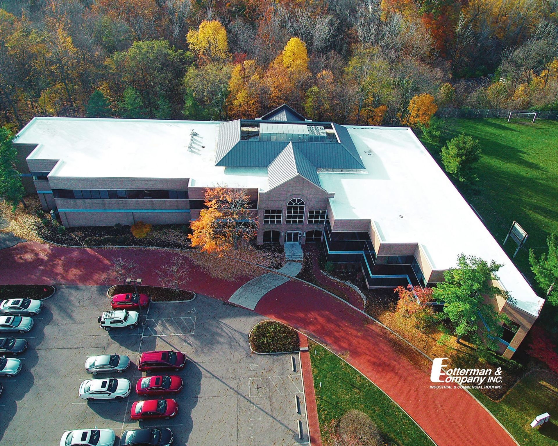 Aerial view of building with red brick entrance and parking lot, surrounded by fall trees.