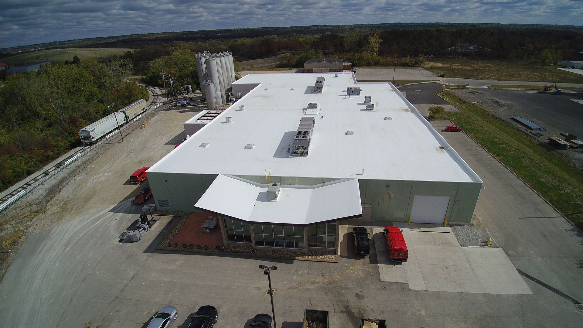 Aerial view of industrial building with white roof and a tall silo. Cars and trucks in the parking lot.