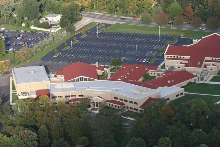 Aerial view of a large beige building complex with red roofs, a large parking lot, and trees.