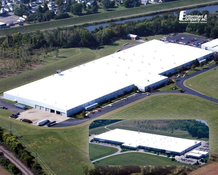 Aerial view of a large white industrial building with a flat roof, surrounded by green grass, trees, and a road.