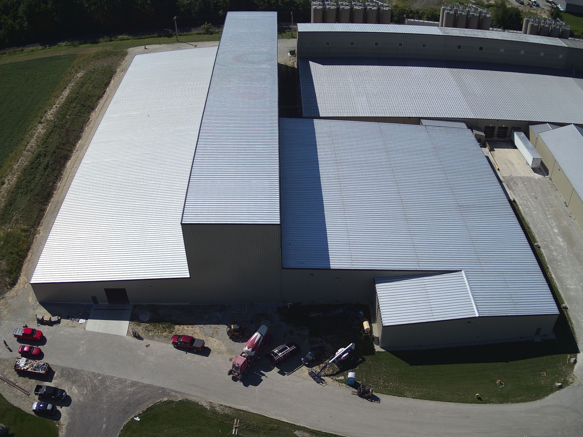 Aerial view of a large industrial building with a corrugated metal roof. A few vehicles are parked in front.