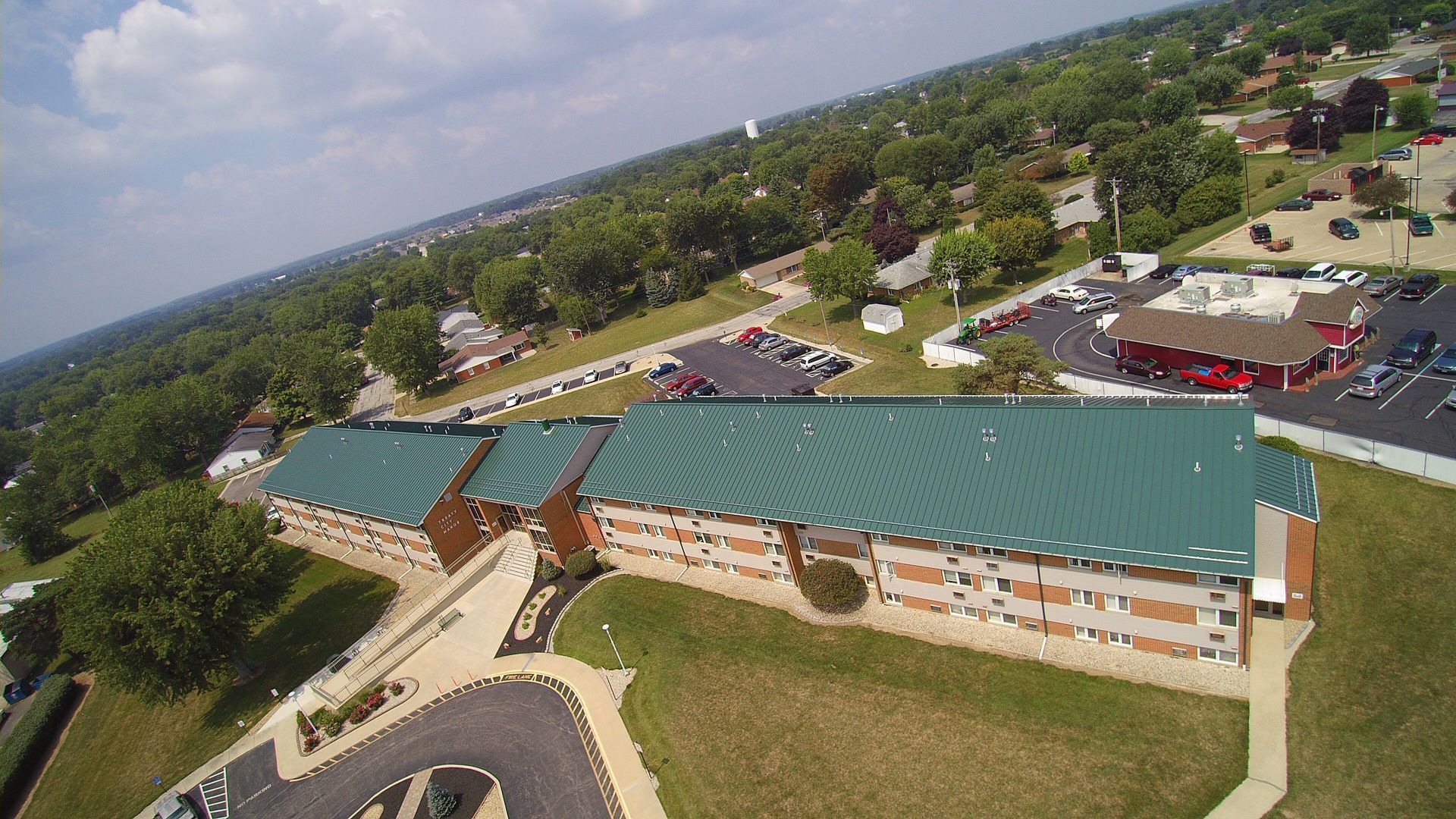 Aerial view of a long, low, brick building with a green roof, possibly a school, surrounded by trees and a parking lot.