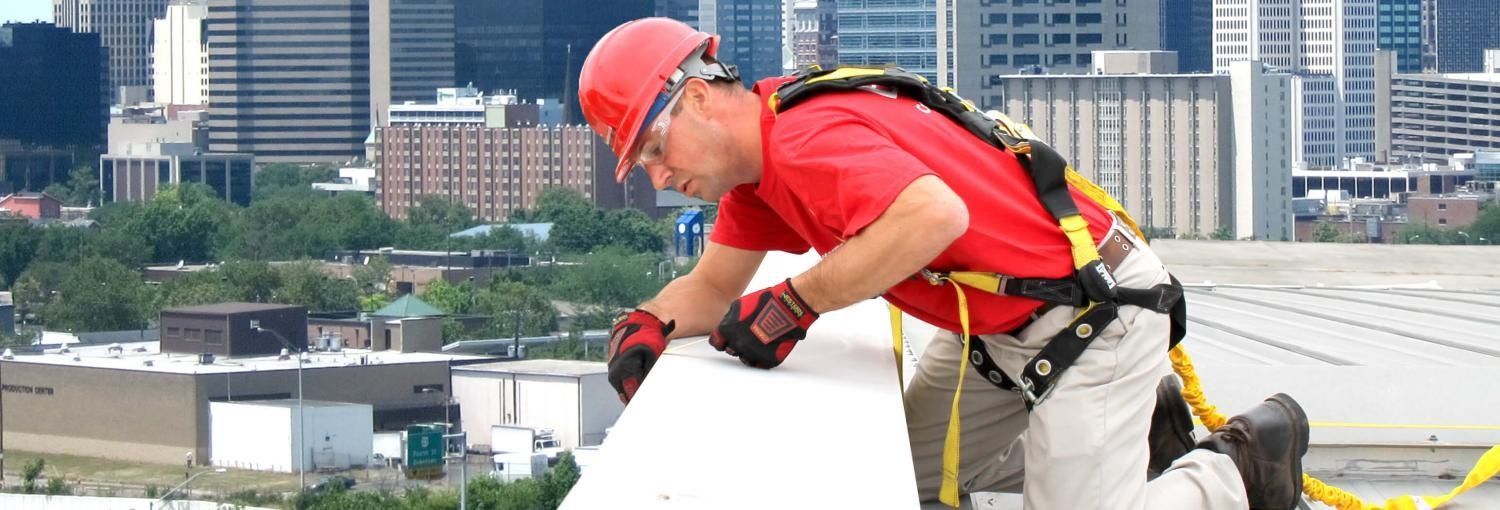 Construction worker wearing safety gear working on a roof with a city skyline in the background.