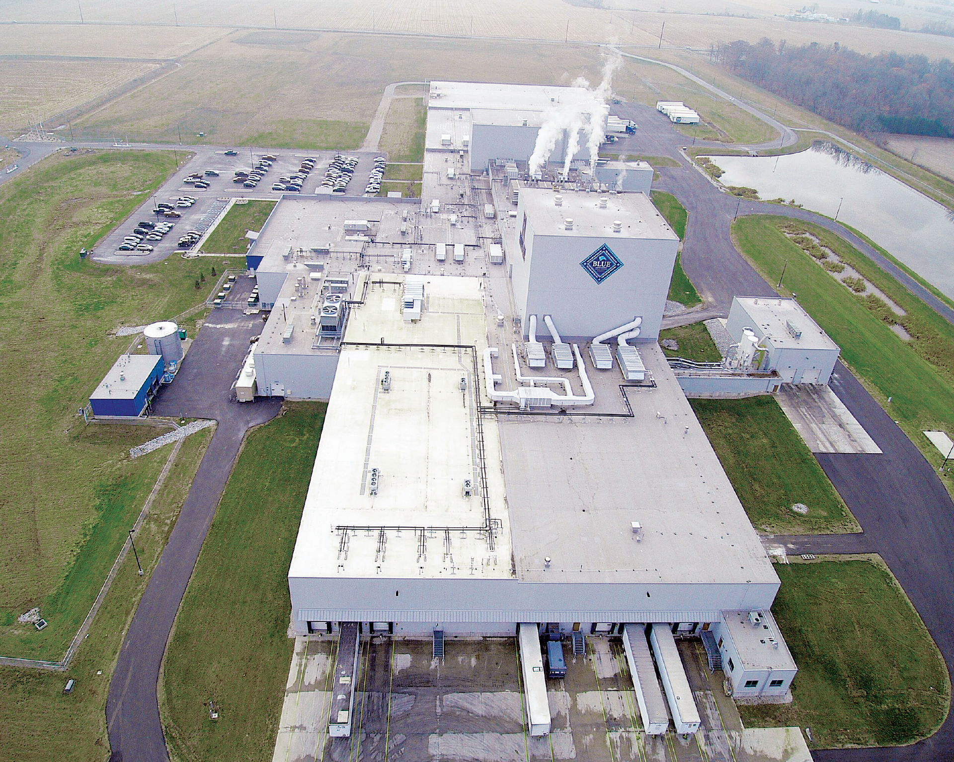 Aerial view of a large industrial factory with multiple buildings, smoke stacks, a parking lot, and a pond.