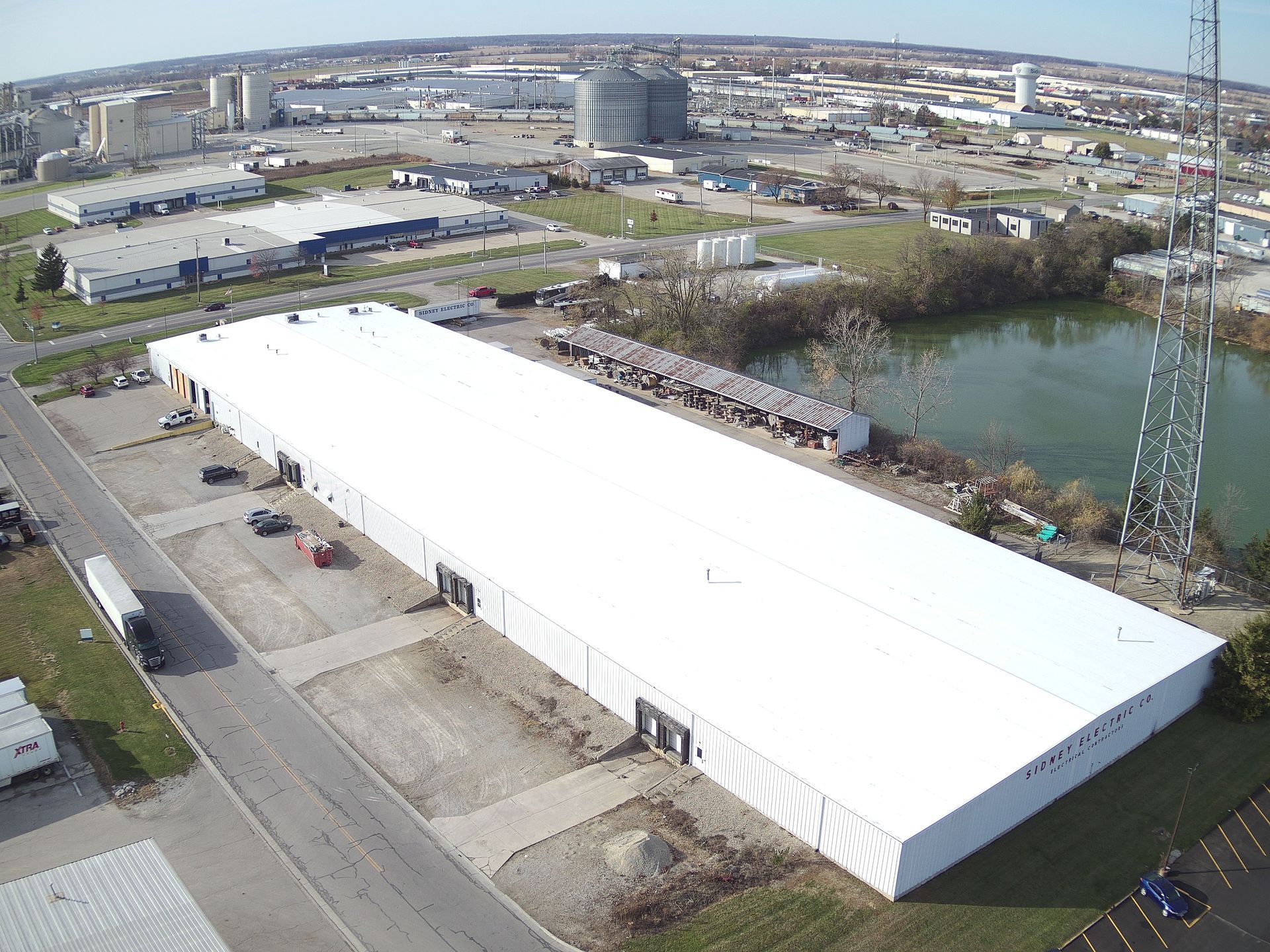 Aerial view of a long, white industrial building with a large parking lot, and surrounding buildings in the background.