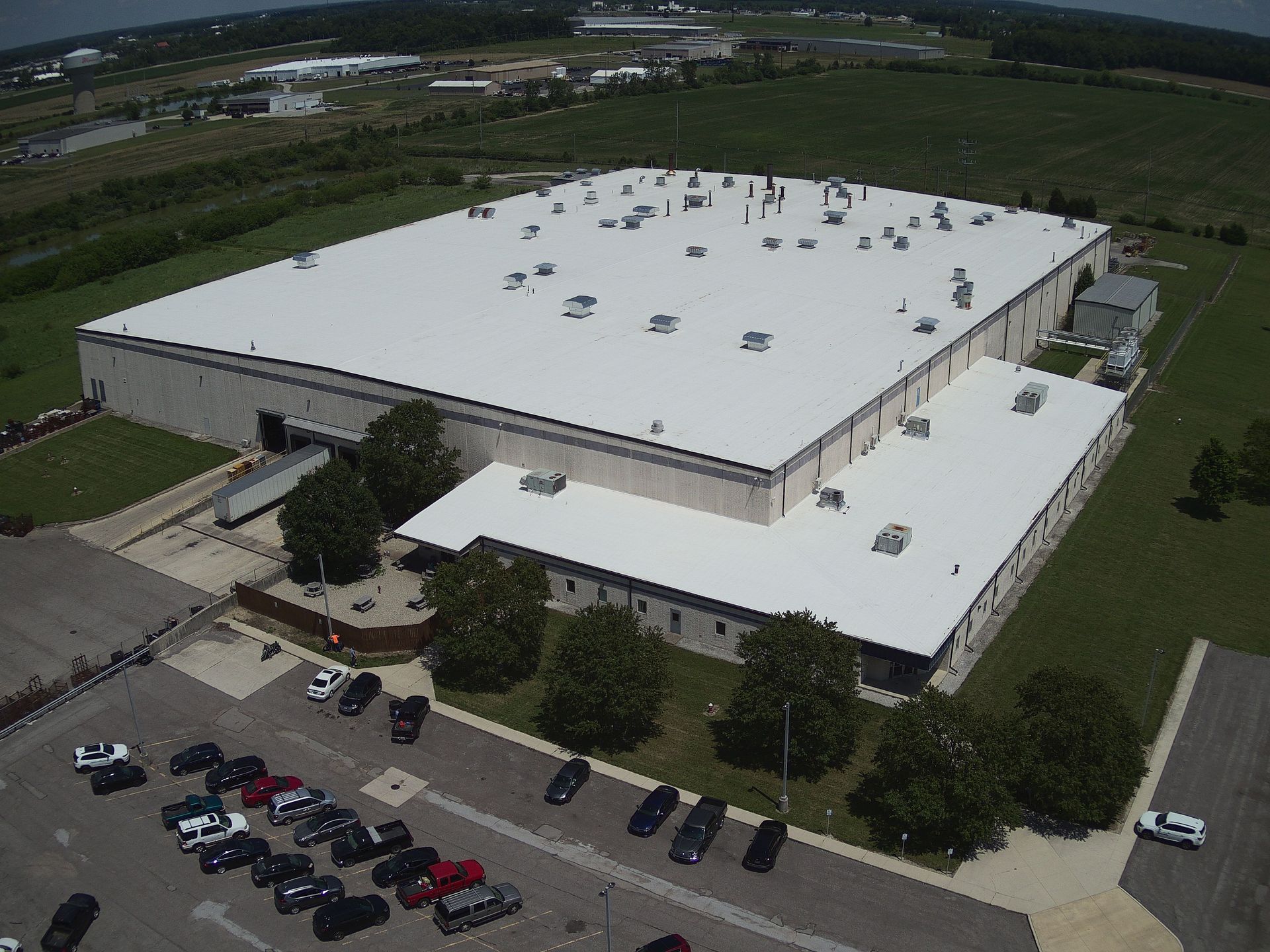 Large industrial building with white roof, parking lot, and surrounding greenery.