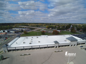 Aerial view of a commercial building with a bright white roof and surrounding parking lot and buildings under a cloudy sky.