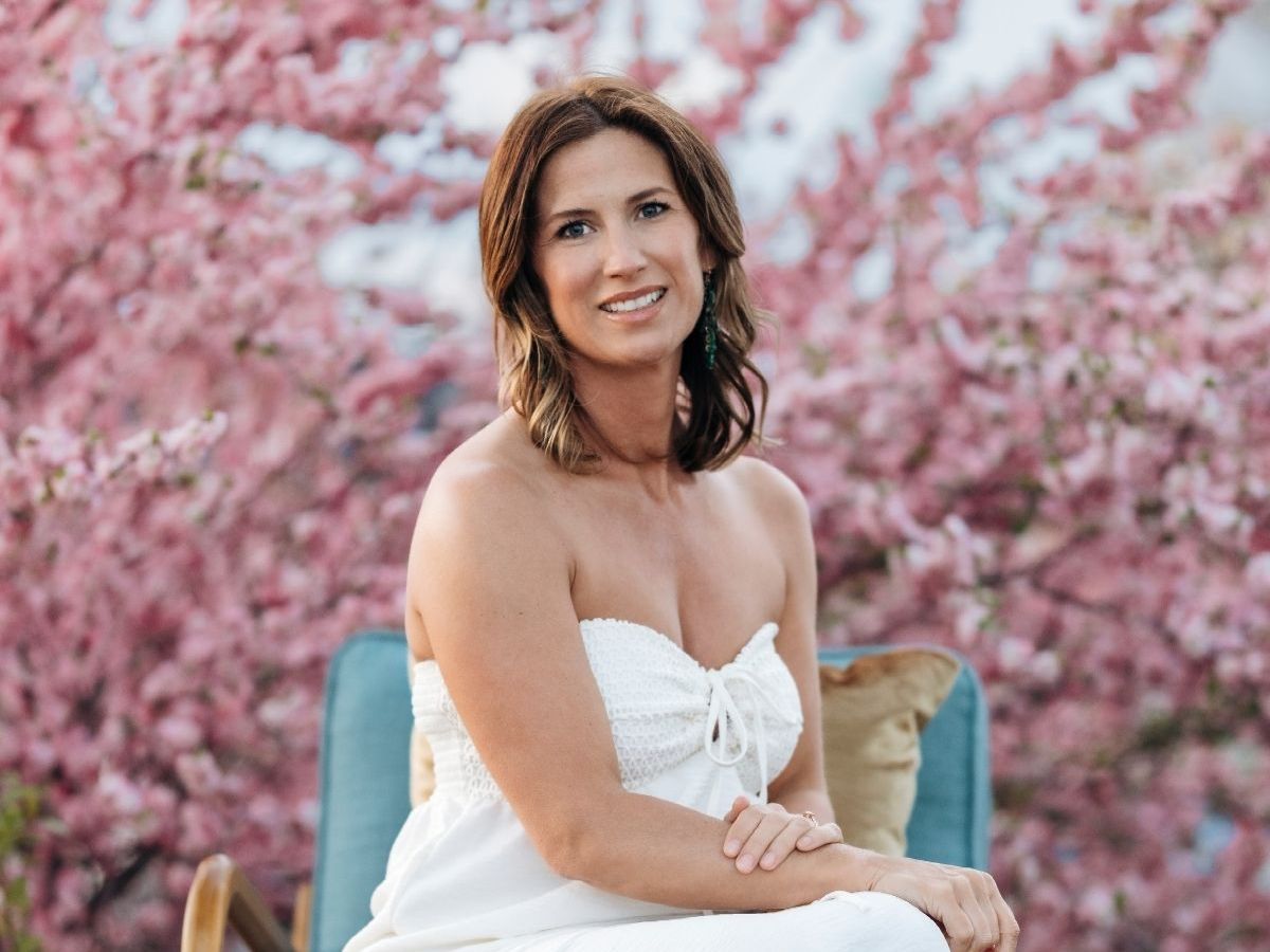 Woman in a white strapless dress sits in a blue chair, smiling, with pink flowers in the background.