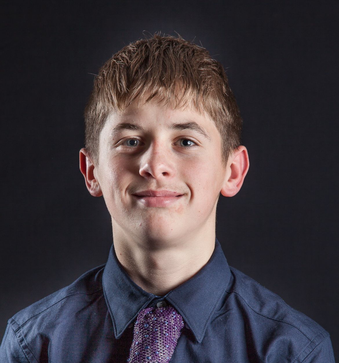 A young man wearing a blue shirt and purple tie smiles for the camera