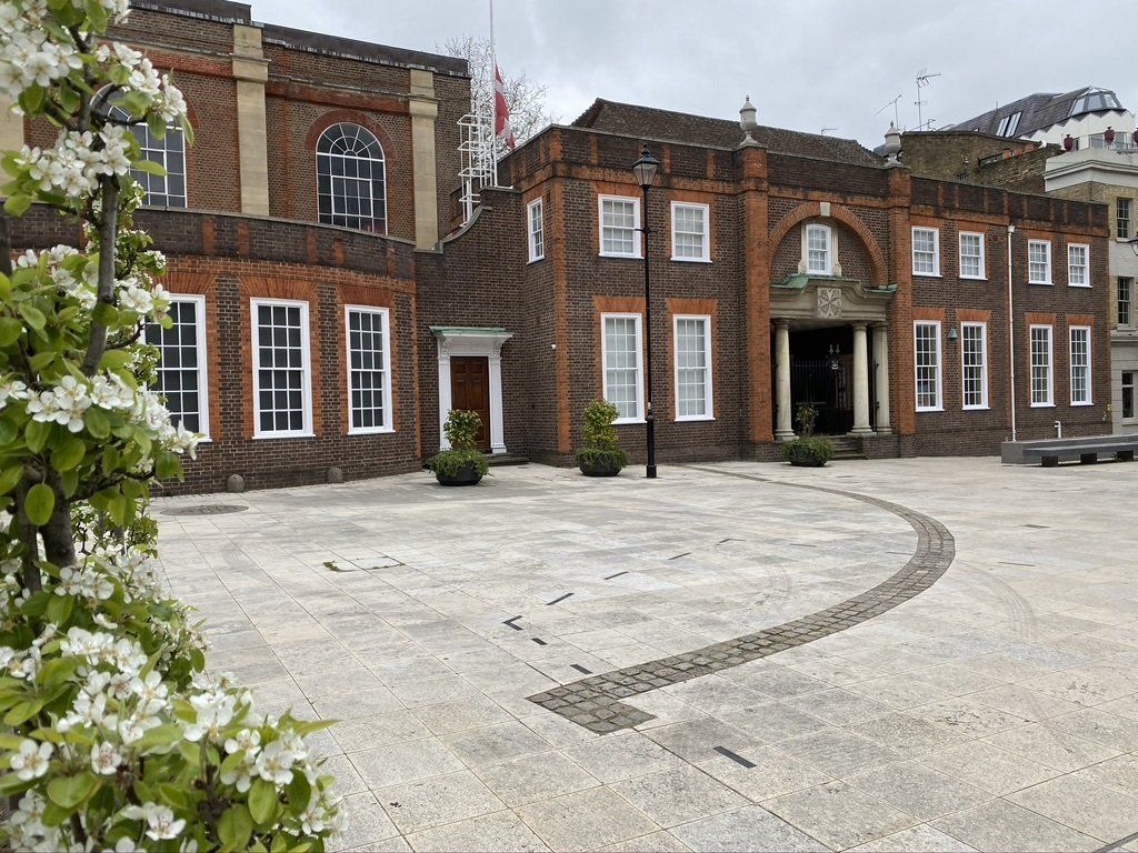 Brick building with columns and a paved plaza, with white flowers in the foreground.