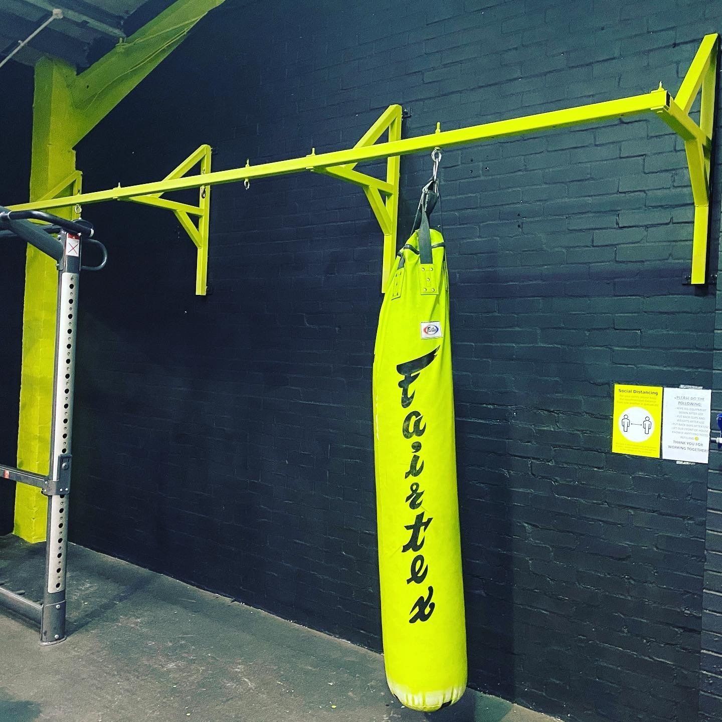 Yellow punching bag hanging from a yellow metal bar on a black brick wall.