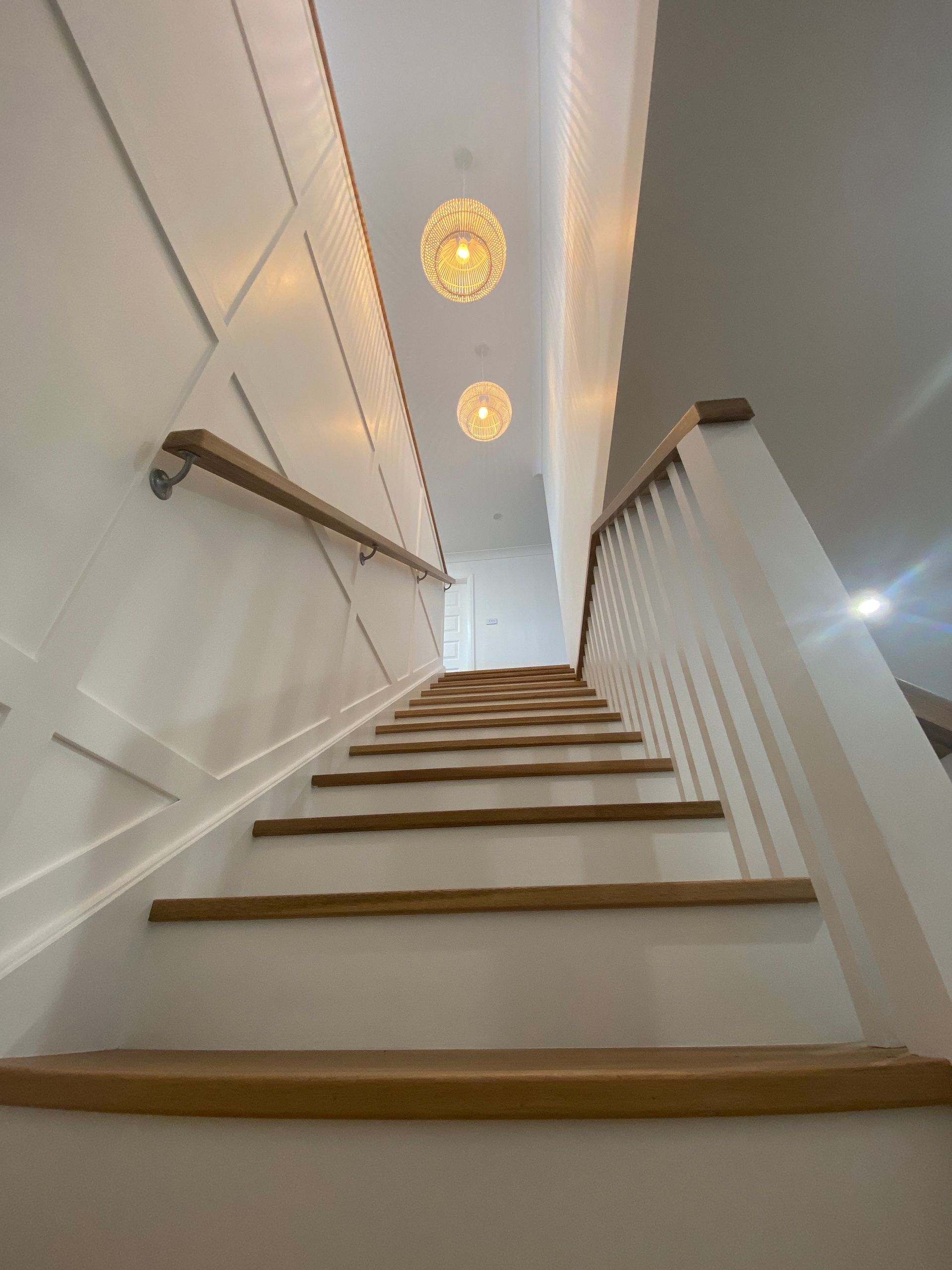 Staircase leading upwards, with wooden steps, white walls, and two decorative lights hanging from the ceiling.