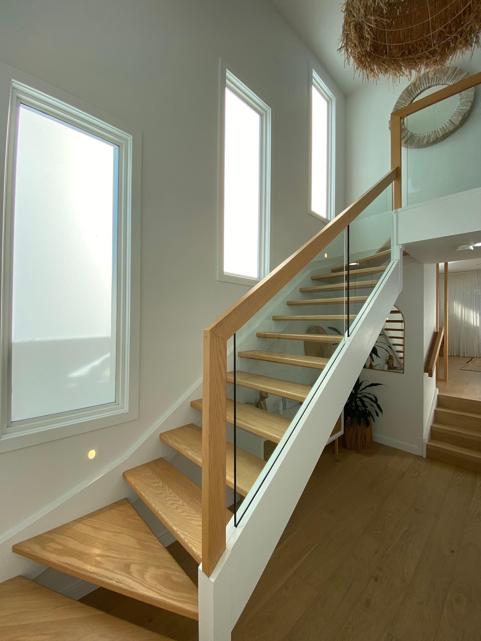 Wooden staircase with glass railing and three tall windows, light-filled interior.