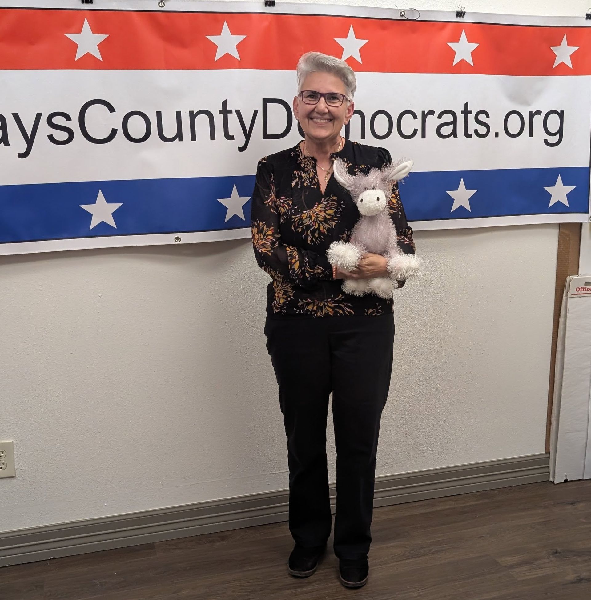 Woman holding a stuffed donkey, standing in front of a banner for the Clay County Democrats.