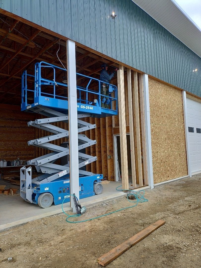 Construction worker on a lift installs wall siding on a commercial building; blue lift, plywood, and green metal siding.