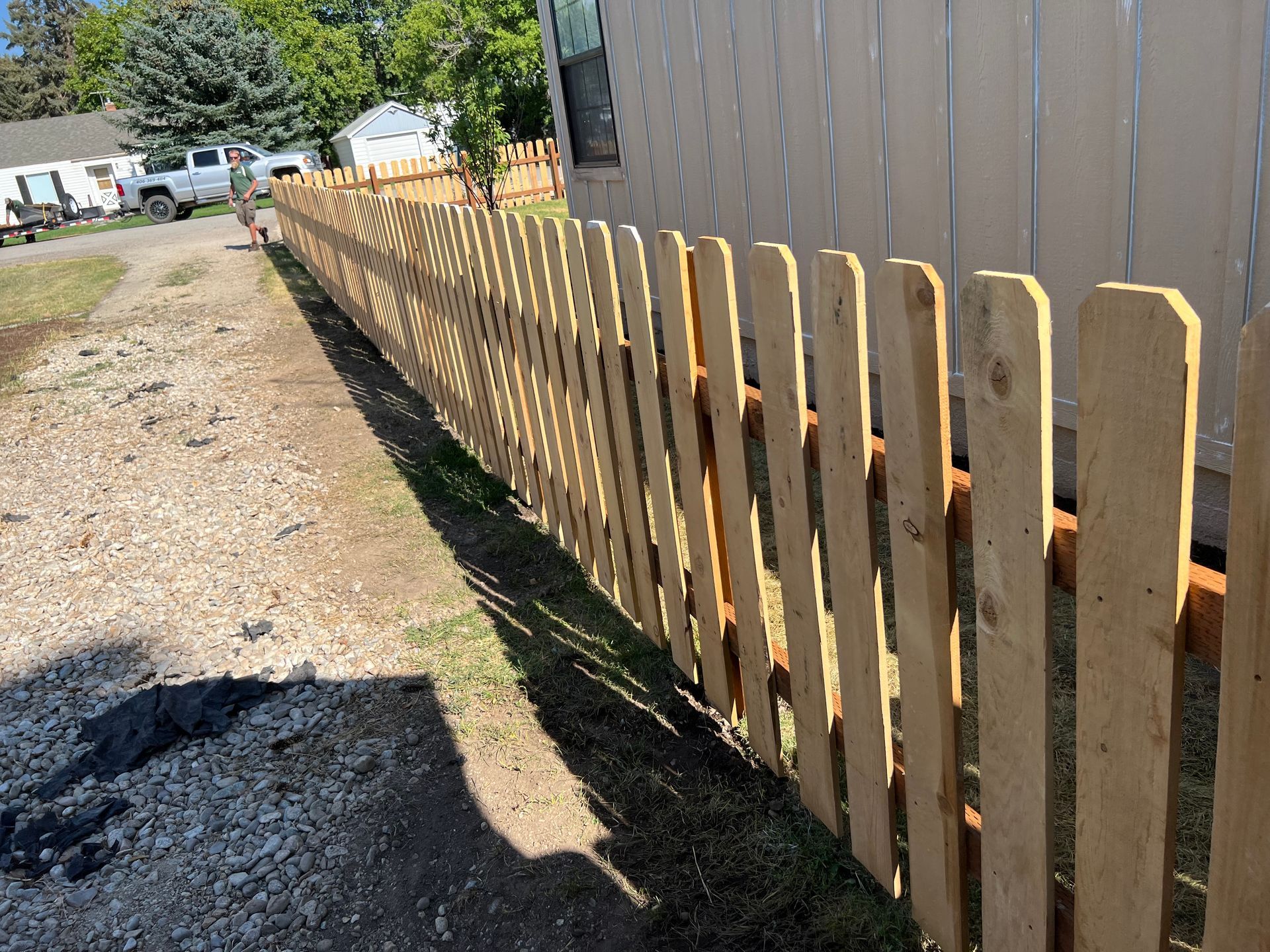 Wooden picket fence along a gravel path and grassy area, beside a beige building on a sunny day.