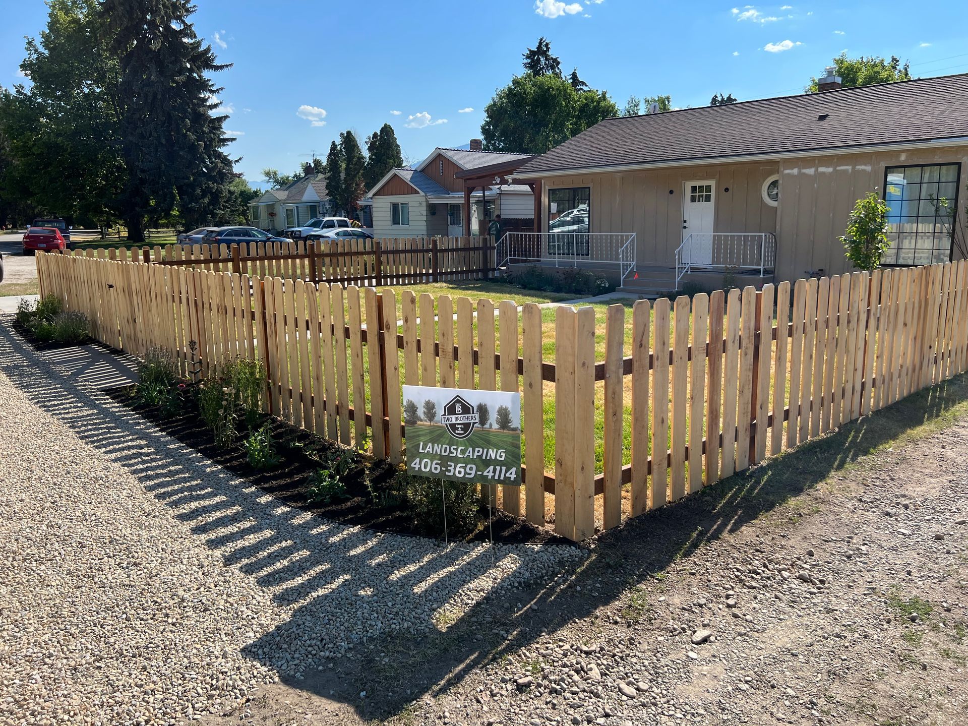 Wooden fence surrounds a small yard, with a sign and landscaping. A tan house is in the background.