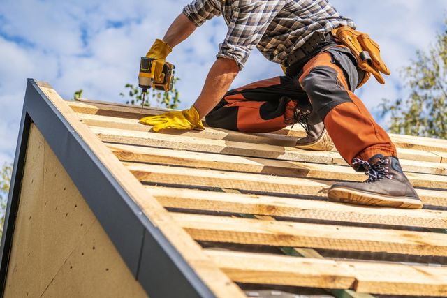 A construction worker in work clothes and yellow gloves uses a power drill to install wooden planks on a sloped roof.