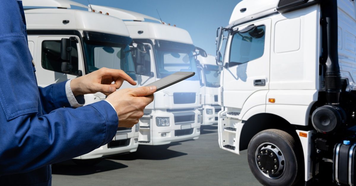 A focus shot of a worker's hands holding a tablet while standing in front of rows of white fleet trucks.