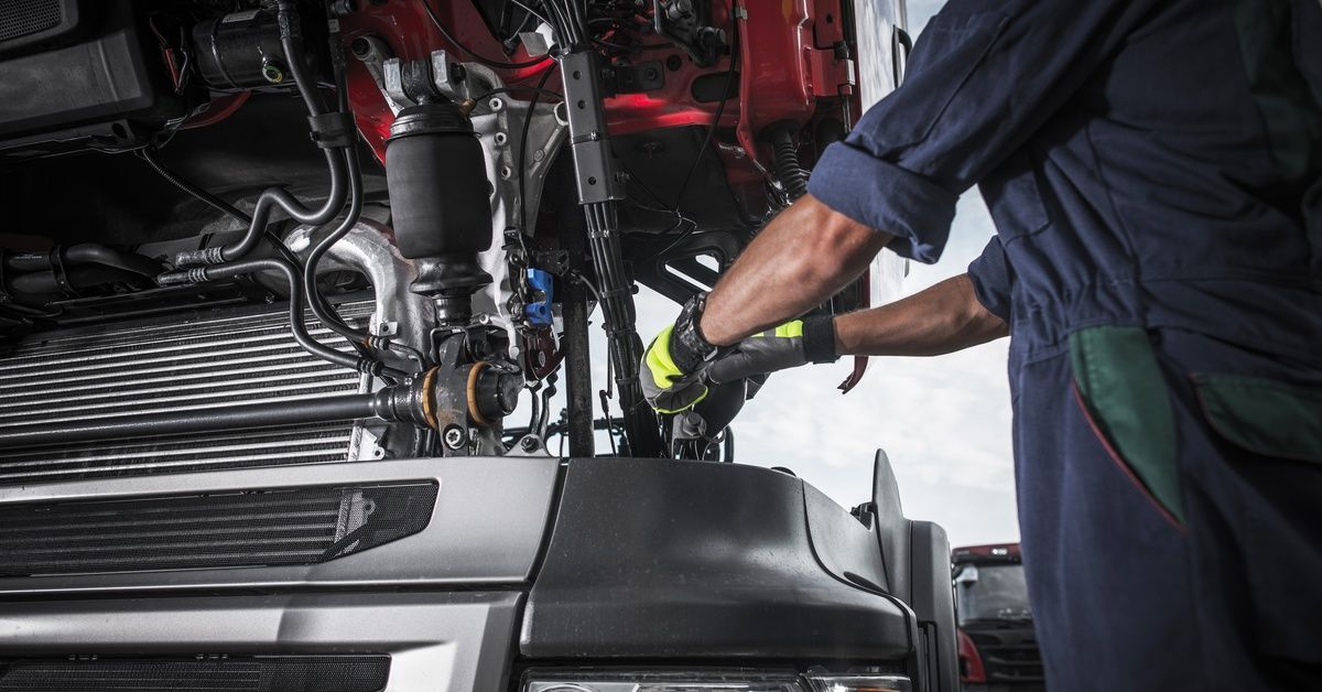 A worker in full mechanic overalls, sunglasses, and a cap, working on the exposed engine of a red truck cabin.