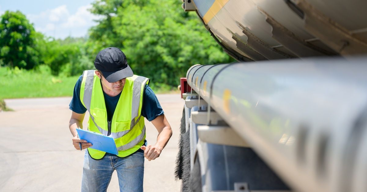 A man in a yellow reflective safety vest bends forward to inspect the tires of a tanker trailer in a parking lot.