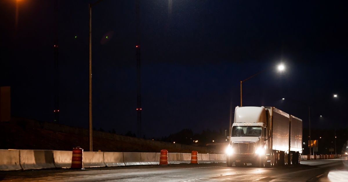 A white semitrailer driving down a freeway at night with its headlights on. It passes by orange traffic drums on the shoulder.