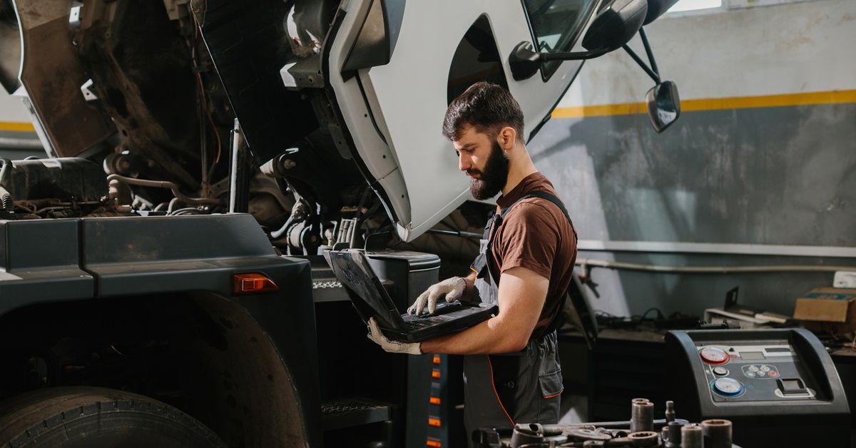 A mechanic holding and referencing a laptop as he stands next to a semitruck with its cab lifted up in a garage.