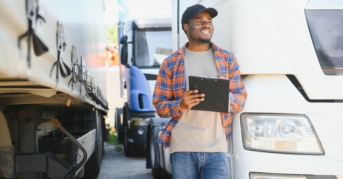 A smiling man walking around a white semi-truck with a clipboard in his hand. He wears a baseball ca