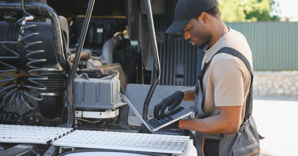 A repair technician in overalls stands in front of the bed of a semitruck, checking something on his laptop.