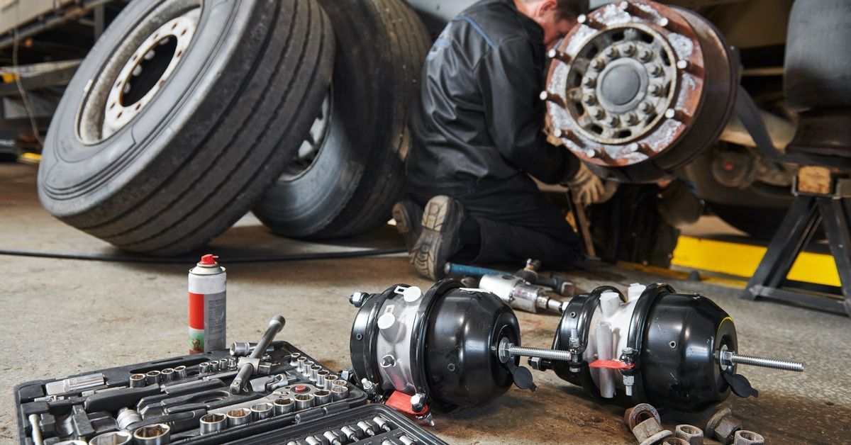 A mechanic works on the exposed wheel hub of a large truck. Spare tires, tools, and brake components lie on the shop floor nearby.