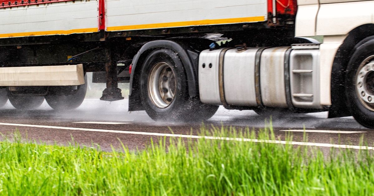 A side view of a semitruck driving on a wet highway, water spraying from the tires beside green grass.