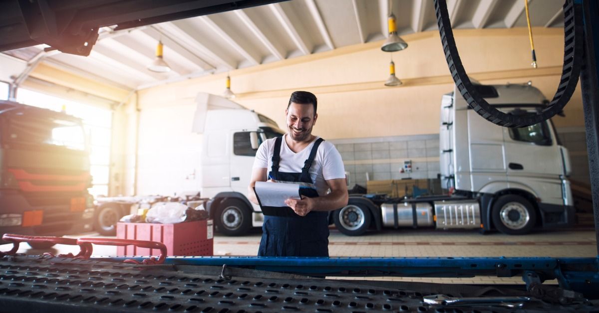 A smiling mechanic stands in a truck repair garage in front of semi-trucks parked in the background.