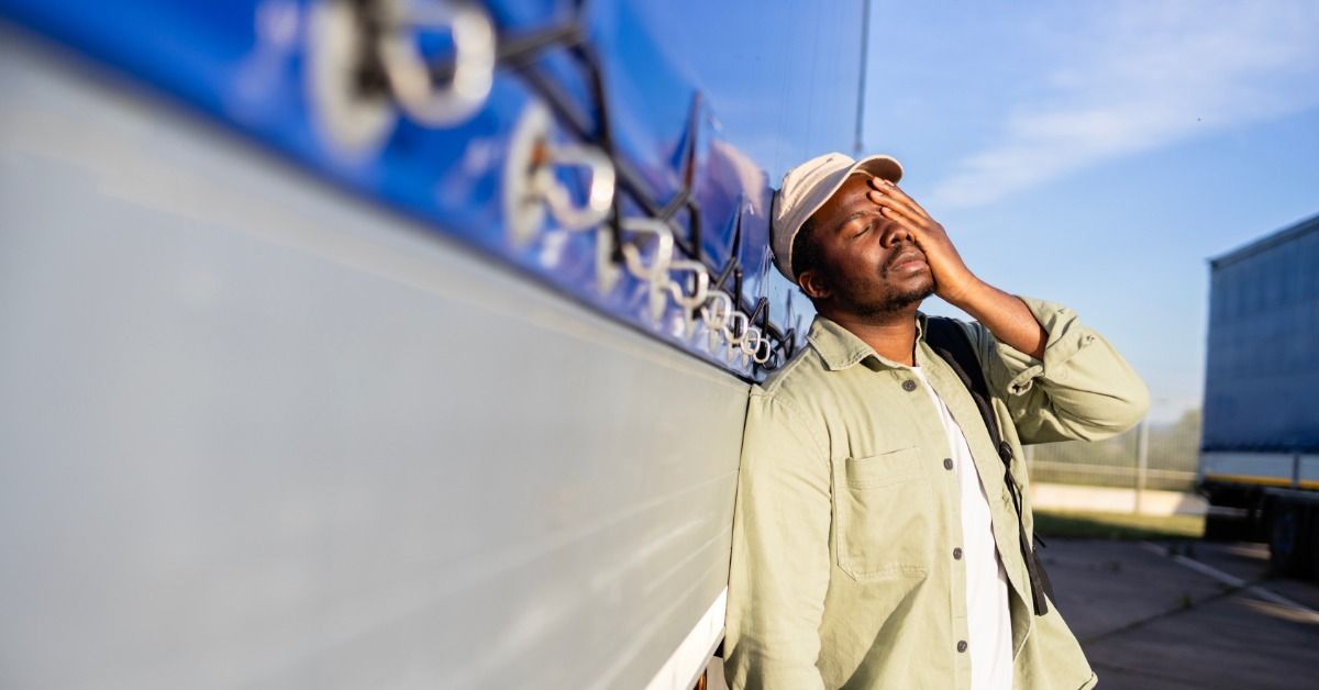 A defeated truck driver leans against a semitrailer with a blue tarp covering, placing a hand over half of his face.