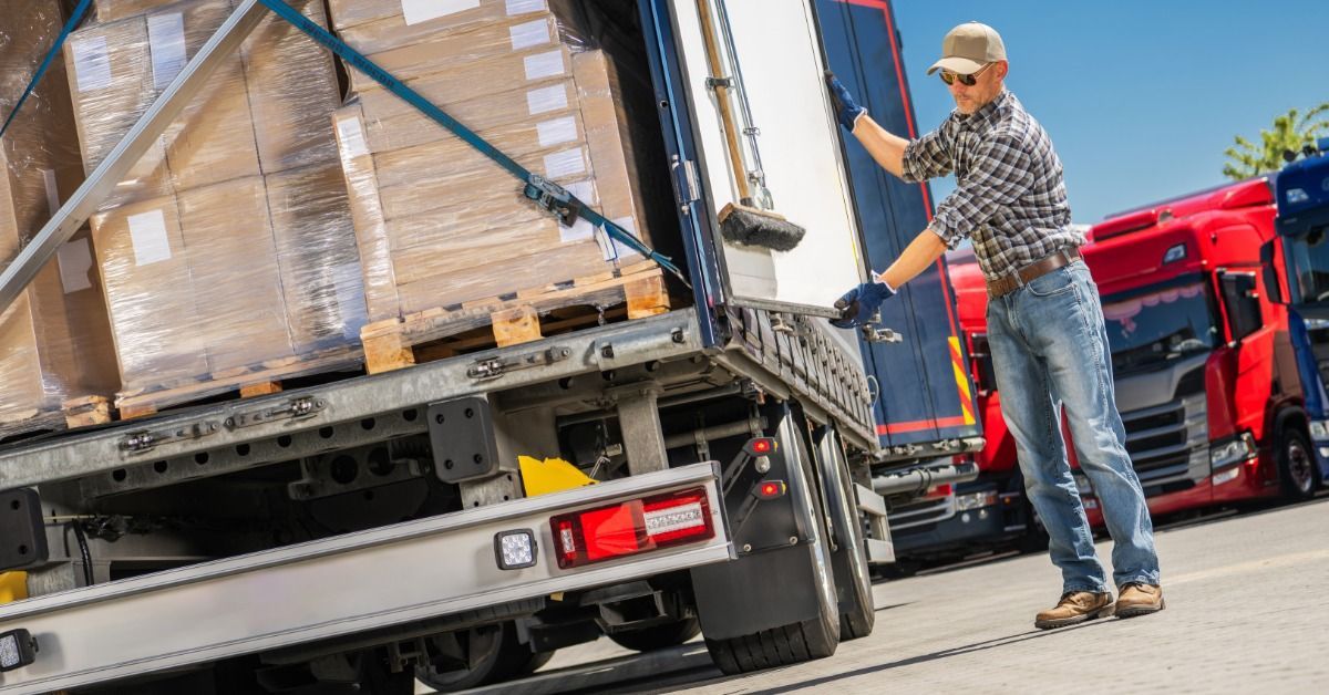 A trucker closes the trailer door to a semitruck fully loaded with palletized boxes that are strappe