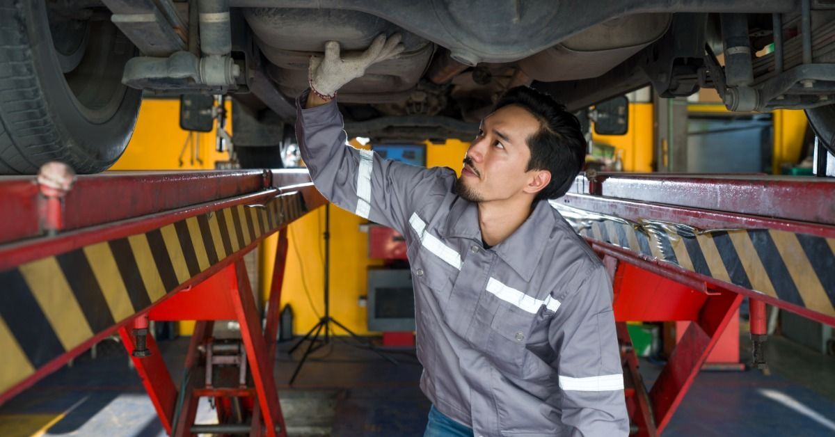 A mechanic under a semitrailer on a lift inspects the undercarriage, reaching up with a gloved hand toward the axles.